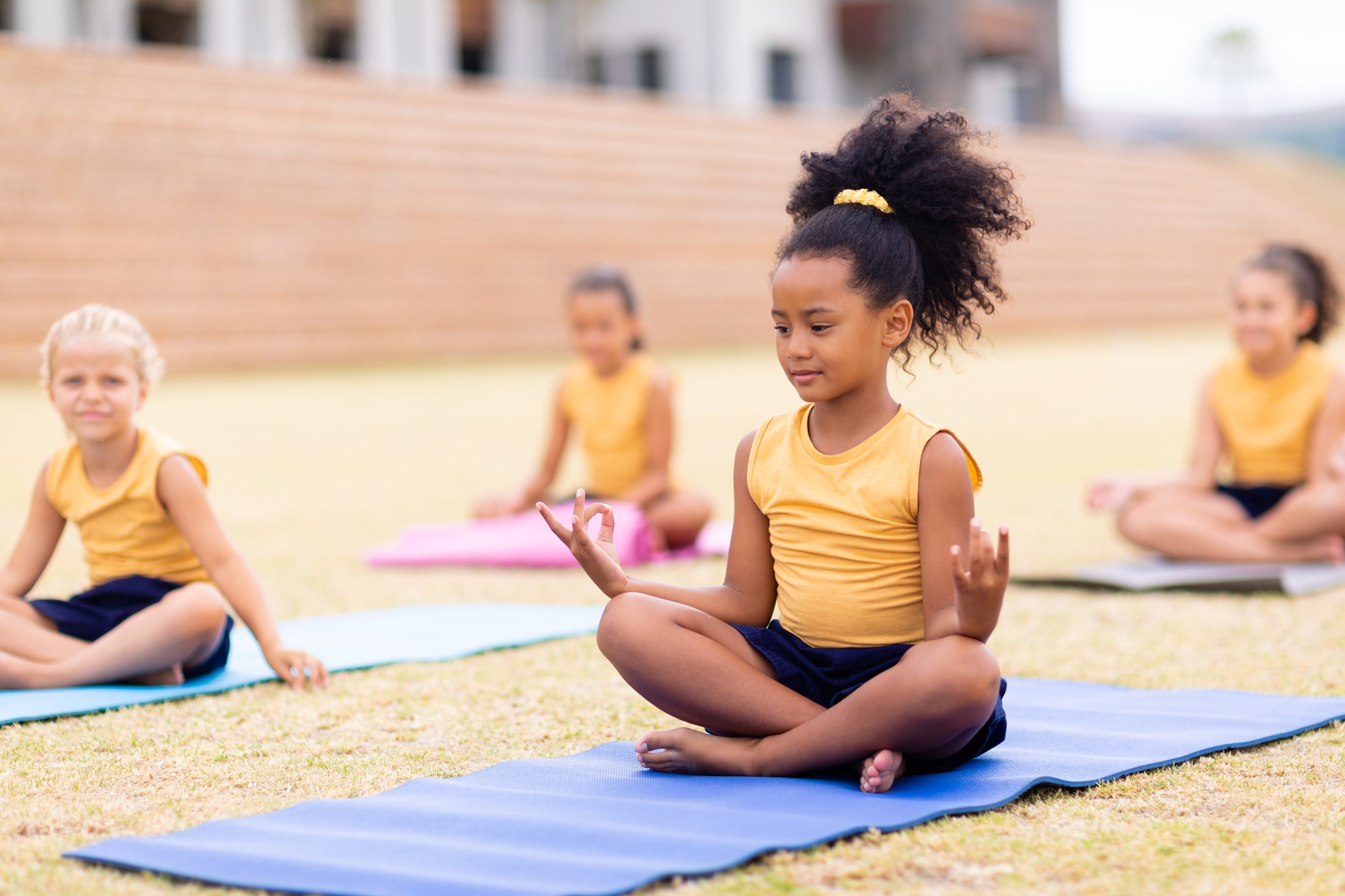Multiracial elementary schoolgirls with gesturing while sitting on yoga mat at school ground